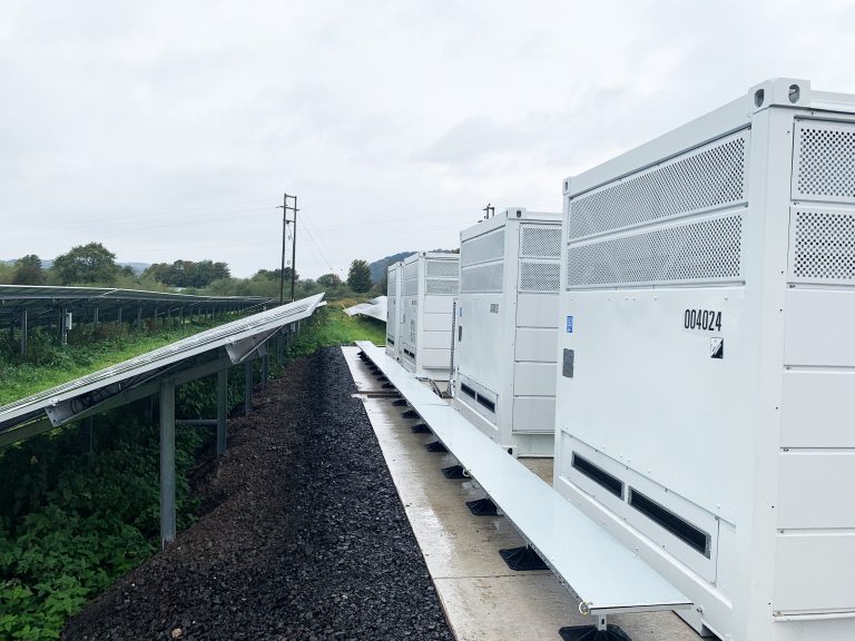 Blue coloured solar panels in a grassy field and white generators sitting behind the panels.