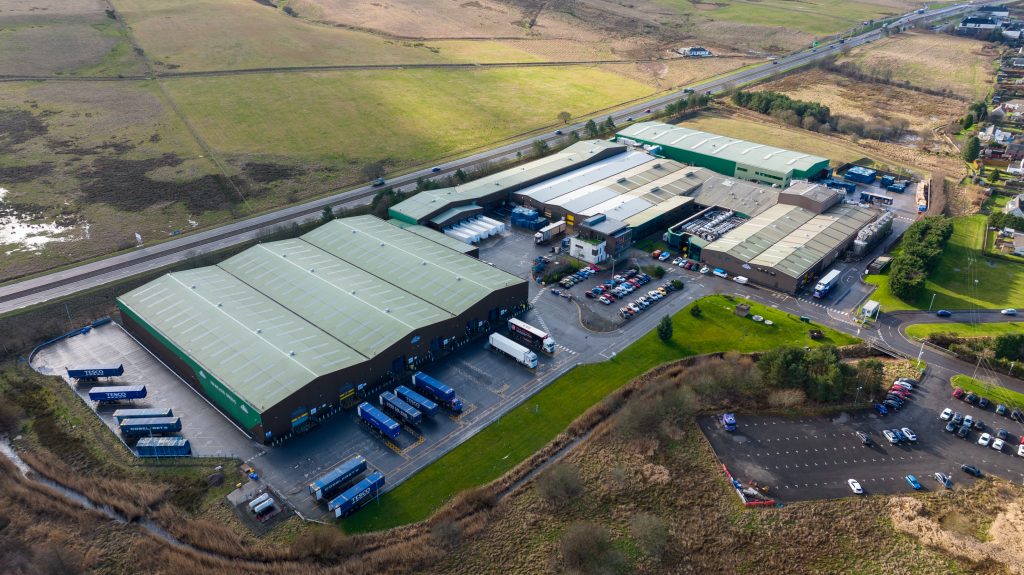 Drone image of large green coloured warehouses, car parking and loading bays for lorries.