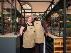 Two middle aged people wearing light brown aprons and dark coloured trousers.